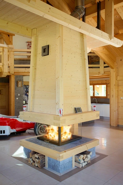 Intérieur chalet bois, grande pièce à vivre avec cheminée centrale suspendue à foyer ouvert 3 faces, habillage bois massif clair, base en pierre naturelle avec niche à bûches