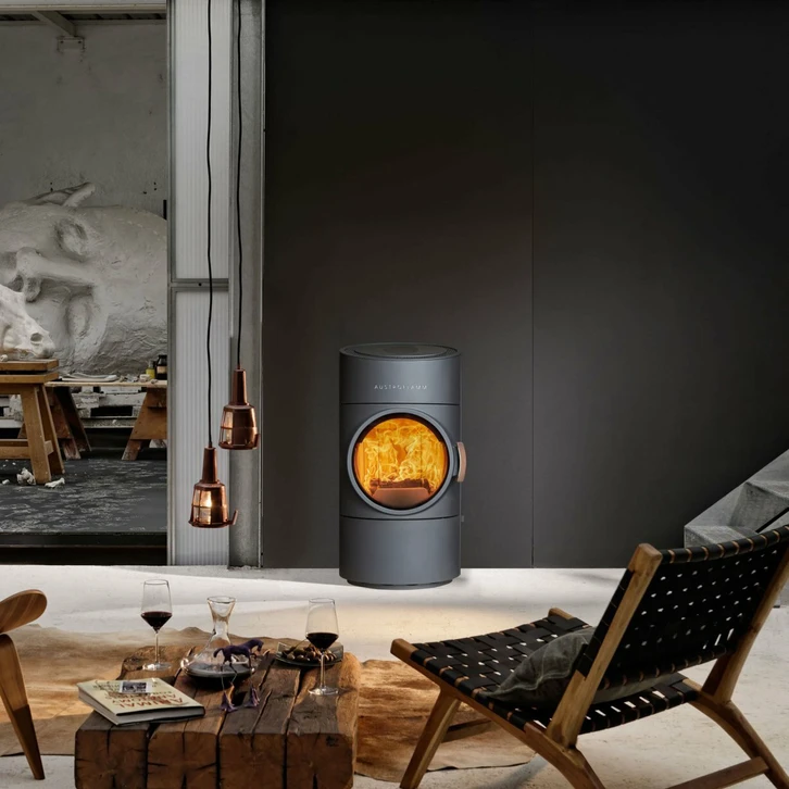 Intérieur moderne avec poêle à bois cylindrique gris anthracite sur pied, flammes vives visibles à travers la vitre panoramique